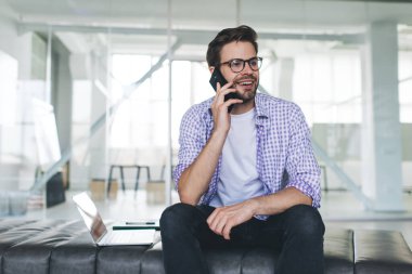 Gülümseyen bin yıllık beyaz iş adamı akıllı telefondan konuşuyor. Modern başarılı adam kavramı. Deri bankta oturan gözlüklü yakışıklı ve açık uzay ofisinde dizüstü bilgisayarı olan genç adam.