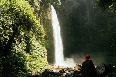 Gündelik giysileri ve şapkasıyla taşlık yolda duran ve Bali 'deki şelalenin görkemli manzarasına hayran olan tanınmamış erkek yolcuların arka planı.