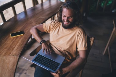 Yüksek açılı genç sakallı erkek serbest çalışan ahşap tezgahta akıllı telefonlu, dizüstü bilgisayarlı ve otel odasının balkonunda çalışırken ekrana bakan.