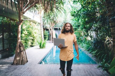 Genç bir turist kameraya bakıyor ve elinde dizüstü bilgisayarla havuzun asfaltlı tarafında yürüyor.