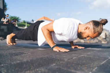 Güneşli bir günde dışarıda antrenman yaparken spor kıyafetleriyle yoga tahtası egzersizleri yapan konsantre bir sporcunun yan görüntüsü.