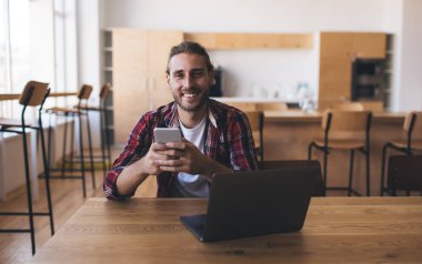 Milenyum Kafkasyalı akıllı telefon kullanıyor ve iş sırasında kameraya bakıyor. Modern başarılı adam kavramı. Genç gülümseyen erkek girişimci açık uzay ofisindeki ahşap masada oturuyor.