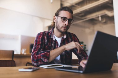 Milenyum Kafkasyalı iş sırasında laptoptan bir şey izliyor. Modern başarılı adam kavramı. Gözlüklü genç girişimci açık uzay ofisindeki ahşap masada oturuyor.