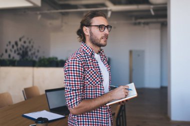 Defteriyle iş sırasında bir şeyler düşünen bin yıllık beyaz iş adamı. Modern başarılı adam kavramı. Gözlüklü genç girişimci açık uzay ofisindeki ahşap masada duruyor.
