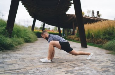 Spor giyimli, sakallı ve spor ayakkabılı güçlü bir erkek sporcunun yan görünüşü. Tahta köprünün altında elleri ve ayakları üzerinde öne eğilip esniyor.
