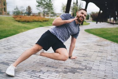 Bulutlu bir günde şehir parkında kaldırımlı yolda antrenman yaparken bacaklarıyla yan tahtayla oynayan sakallı bir erkek sporcu.