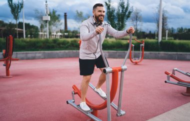 Spor sahasında bulutlu gökyüzüne karşı spor yaparken gülümseyen ve egzersiz yapan sakallı bir erkek sporcu.