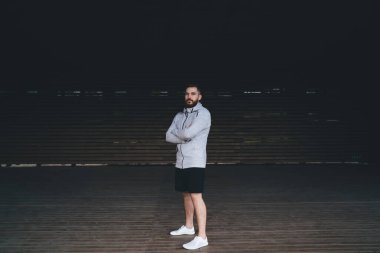 Spor giyimli, kendinden emin bir erkek sporcu. Ahşap bir platformda duruyor. Kollarını kavuşturup kameraya bakıyor.