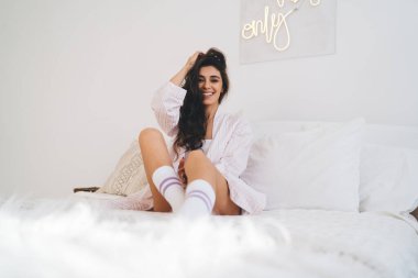 Pijamalı genç bir kadın uzun saçlara dokunuyor ve hafta sonunu rahat bir yatak odasında kameraya bakarak geçirirken yatakta oturuyor.