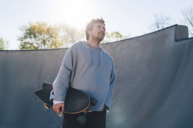 Alçak açılı mutlu kaykaycı erkek kaykaycı kaykay parkında durup özgürlüğün ve üretken eğitimin tadını çıkarıyor.