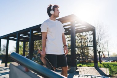 Sıradan giyinen erkek kaykaycı, hafta sonu kaykay parkında gözlerini kaçırırken kulaklıkla şarkı söylerken eğleniyor.