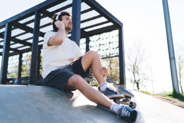Tişörtlü ve şortlu neşeli genç kaykaycı kaykaylı beton rampada oturmuş kulaklıkla müzik listesinden şarkılar dinliyor.