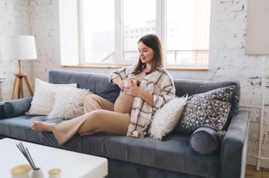 Pozitif kadın, sıcak oturma odasında dinlenirken rahat bir kanepeye oturup, pencereye yaslanıp sıcak içeceğin keyfini çıkarıyor.