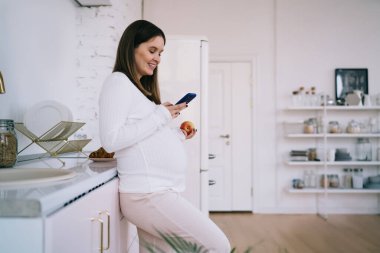 Modern mutfakta elma yerken, günlük kıyafetlerini giymiş, cep telefonuyla sohbet eden ve doğum izni sırasında boş vakitleri olan genç bir kadının yan görüntüsü.