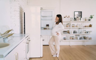 Düşünceli, genç ve hamile bir bayanın mutfakta parmakları dudaklarında durması ve buzdolabından gerekli eşyalar bulup evde vakit geçirmeyi planlarken atıştırmalık bir şeyler hazırlaması.