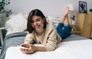 Uzun boylu, iyimser, yalın ayaklı, koyu saçlı, balıkçı yaka yakalı, koyu renk saçlı, siyah saçlı, akıllı telefondan mesaj atarak yatakta uzanan, gülümseyen ve kameraya bakan bir kadın.