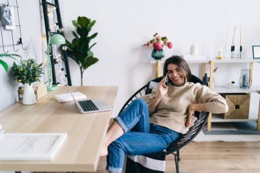 Telefon görüşmeleri sırasında sandalyede dinlenen ve evdeki oturma odasında dizüstü bilgisayarla çalışırken kameraya bakan gündelik kıyafetlerle gülümseyen kadın girişimci.