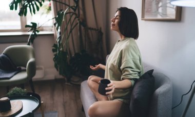 Kahve fincanı ile meditasyon yapan genç bir kadın. Gözleri kapalı. Oturma odasındaki kanepede rahatça oturuyor.