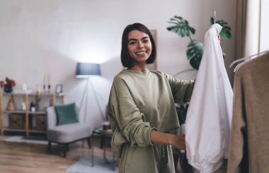 Modern oturma odasında beyaz elbiseler giyerken gülümseyen ve kameraya bakan gündelik kıyafetli İspanyol bir kadın.