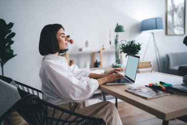 Yoğunlaştırılmış serbest çalışan, günlük giysiler içinde dizüstü bilgisayarla masada oturan ve evdeki uzak projeler üzerinde çalışan kadın.