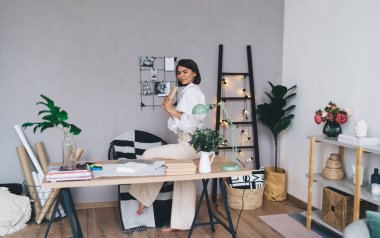 Beyaz tişörtlü, bej pantolonlu, serbest çalışan, elinde kitapla masa başında oturan, yaratıcı çalışma alanında çalışan ve kameraya bakan bir adam.