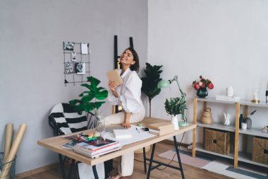 Gülümseyen, başı arkaya eğik, gözleri kapalı, elinde not defteri olan ve yaratıcı çalışma alanında çalışırken masasında oturan serbest çalışan genç bir bayan.