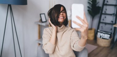 Evdeki rahat koltukta otururken cep telefonunda selfie çekerken bej balıkçı yaka yakalı, parlak gözleri olan ve saçlarına dokunan neşeli, genç ve şık bir bayan blog yazarı.