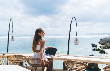 Yazlık kıyafetiyle gülümseyen, uzun saçlı, hasır koltukta dizüstü bilgisayarla oturan ve uzak bir projede çalışırken okyanus manzarasının tadını çıkaran genç bir kadın.