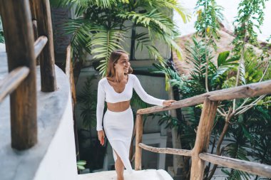 Beyaz tişörtlü, etekli, gülümseyen kadın serbest yazar yaz tatilinde Tayland 'a seyahat ederken merdivenlerden çıkıp doğaya hayranlık duyuyor.