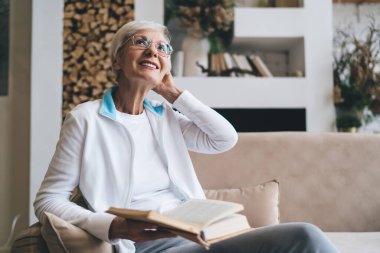 Gözlüklü, gülümseyen ve başını çeviren, haftasonu evde oturup kitap okuyan yaşlı bir kadın.