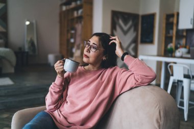 Pembe kazaklı yaşlı bir kadın, elleri başının üstünde koltukta dinleniyor. Sıcak içkinin tadını çıkarırken, boş zamanlarının tadını çıkarıyor.