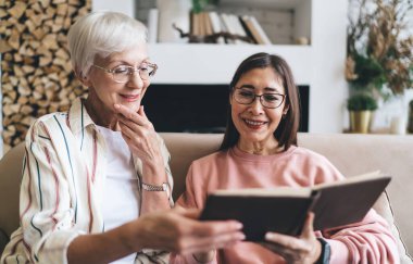 Güncel giysiler ve gözlük takan, fotoğraf albümüne bakan, modern apartmandaki kanepede dinlenirken nostalji yapan pozitif, çok kültürlü kadınlar.
