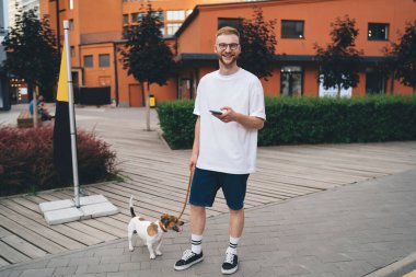 Gözlüklü, beyaz tişörtlü, uzun sakallı genç bir adam tasmalı Jack Russell Terrier köpeği ve akıllı telefonuyla şehir merkezinde gülümseyerek kameraya bakıyor.