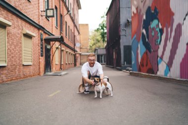 Sıradan giysiler içinde mutlu genç adam şehrin yakınlarındaki asfalt yolda otururken itaatkar Jack Russell Terrier 'ı okşuyor.