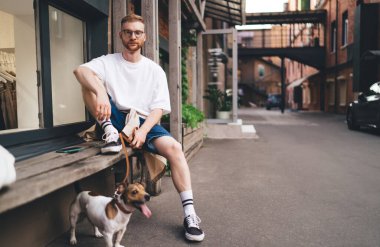 Hafta sonu inşaata karşı ahşap bankta Jack Russell Terrier ile rahat rahat takılan genç bir erkek vücudu.