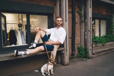 Jack Russell Terrier 'ın yanındaki ahşap bankta oturan ve yürüyüşten sonra başını çeviren düşünceli, genç bir erkek.
