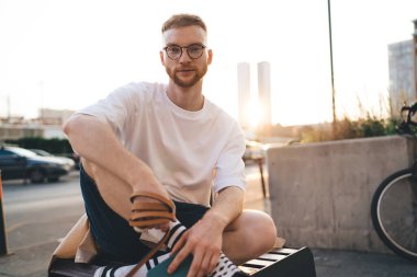 Pozitif sakallı, gözlüklü, bacak bacak üstüne atmış bir şekilde bankta oturan ve hafta sonları yol kenarında dinlenirken kameraya bakan bir erkek.