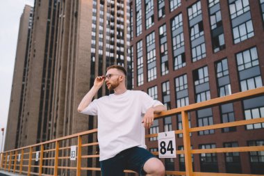 Ciddi, sakallı, sıradan kıyafetli, çatıda duran, parmaklıklara yaslanan, gözlüklere dokunan ve modern, çok katlı binalara bakmayan genç bir adamdan.