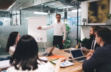 Resmi kıyafetler içindeki genç iş adamları ellerinde kağıtlar ve dizüstü bilgisayarlarla ahşap masada oturuyor ve meslektaşlarının projeyi açıklamalarını dinliyor ve ofisteki ters çizelgeye işaret ediyorlar.