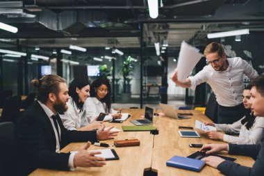 Bir grup farklı iş adamı ellerinde aletlerle oturup birlikte çalışırken, meslektaşları da çağdaş çalışma alanındaki ahşap masaya dökümanları koyuyor.