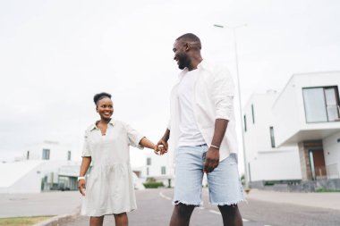 Gülümseyen Afrikalı genç adam ve kadın sıradan kıyafetler içinde birlikte paten kayıyor ve modern evlerin yanındaki asfalt yolda el ele tutuşup birbirlerine bakıyorlardı.