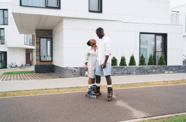 Neşeli siyahi kadın ve erkek, günlük giysiler ve tekerlekli patenler giyip sokakta yan yana durup birbirlerine bakıyorlar ve mutlu bir şekilde gülümsüyorlar.