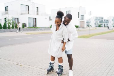 Keyifli Afrikalı Amerikalı erkek arkadaş, hafta sonları şehir sokaklarında kaykay yaparken kız arkadaşına yardım ediyor ve gülümsüyor.