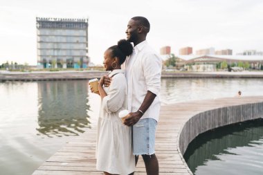 Neşeli Afrikalı Amerikalı erkek, siyah kadını arkadan kucaklarken ahşap köprüde durmuş kahve fincanlarıyla ve parlak bir akşamda gözlerini kaçırıyor.