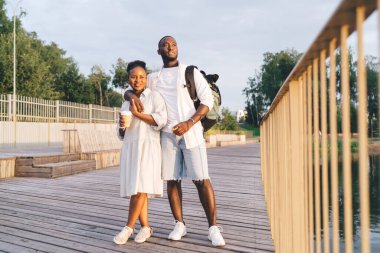 Sırt çantalı Afro-Amerikan bir çift ve ahşap kaldırımda dikilip kafalarını çevirerek kahve içiyorlar.