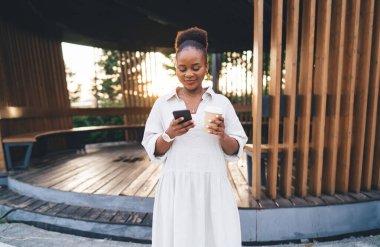 Genç Afro-Amerikalı kadın gündelik giysiler içinde elinde kahve bardağıyla ahşap oyuğun yanında duruyor ve hafta sonları şehir parkında cep telefonu kullanıyor.