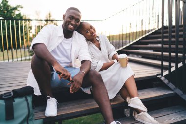 Parktaki tahta merdivenlerde dinlenirken ve kameraya bakarken gülümseyen Afrikalı Amerikalı adamın omzuna yaslanan kağıt bardaklı çekici siyahi kadın.