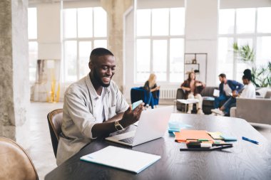 Afrika kökenli Amerikalı girişimci cep telefonu kullanıyor ve ahşap masada otururken gülümsüyor ve modern ofiste dizüstü bilgisayarla iş kurma üzerinde çalışıyor.