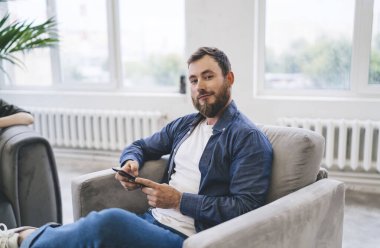 Kendine güvenen sakallı, sıradan kıyafetleri olan, elinde cep telefonuyla koltukta oturan ve ofisteki kameraya bakan bir girişimci.