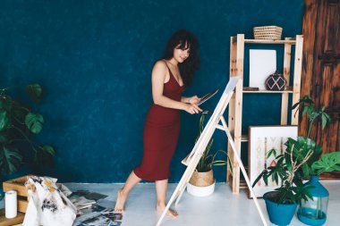 Çıplak ayaklı genç kadın ressamın sehpanın yanında duran ve yeşil bitkilerin yanında boya fırçasıyla tuval üzerinde yaratıcı sanat çalışması yapan.